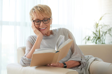 Senior blond woman in glasses resting on sofa and reading a book - Powered by Adobe