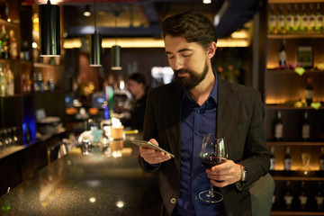 Smiling bearded man drinking red wine in restaurant and texting with friends