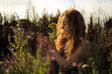 Woman with beautiful cascading long red hair standing amongst wild flowers highlighted by the sun 