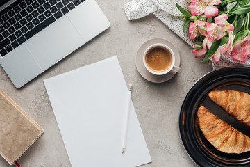 top view of cup of coffee with blank paper, croissants and laptop on concrete surface