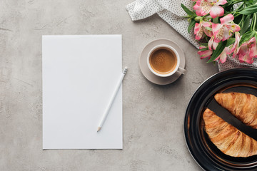 top view of cup of coffee with blank paper, croissants on plate and alstroemeria bouquet on concrete surface