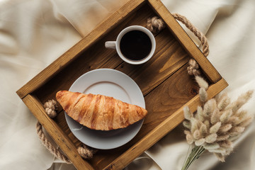 top view of coffee and croissant on tray with lagurus ovatus bouquet on beige cloth, breakfast in...