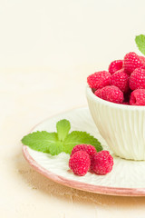 Ripe raspberries with green mint leaves in cup and saucer on pastel yellow background.