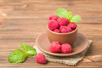 Ripe raspberries with green mint leaves in brown cup and saucer on sackcloth and wooden background.