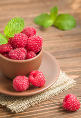 Ripe raspberries with green mint leaves in brown cup and saucer on sackcloth and wooden background.