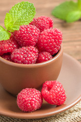 Ripe raspberries with green mint leaves in brown cup and saucer on sackcloth and wooden background.