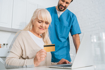 Obraz premium smiling social worker looking at senior woman holding credit card and using laptop