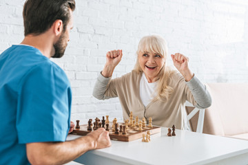 triumphing senior woman celebrating victory and looking at young social worker after chess game