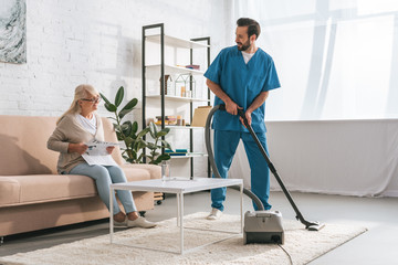 smiling social worker cleaning carpet with vacuum cleaner and looking at senior woman reading...