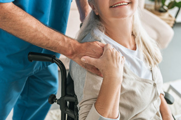 cropped shot of smiling senior woman in wheelchair holding hand of caregiver