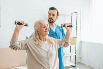 social worker looking at smiling senior woman training with dumbbells