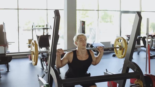 Blonde man holding barbell on the shoulder behind the neck while crouching and doing squats exercise in gym
