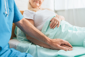 Obraz premium cropped shot of male nurse holding hand of sick senior woman lying in hospital bed
