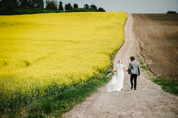 Bride, groom walking on way spring field of yellow rapes flowers, canola field. A newlywed wedding couple running on a country straight road for their honeymoon. Top rear view.