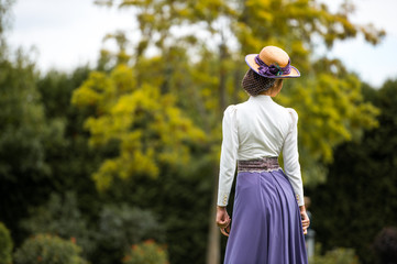 Beautiful girl in a vintage dress. Girl on a blurred background. A girl in a white blouse and a purple dress. A woman is dressed in retro style. The girl is straightening the hat on her head.