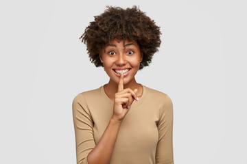 Glad lovely African American female asks to keep silence, holds index finger on lips, has cheerful expression, dressed casually, poses against white background. People, secrecy and ethnicity concept