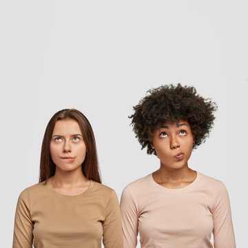 Vertical shot of two confused multiethnic women bite lower lips, looks with puzzlement upwards, dressed casually, try to solve problem, isolated over white background with blank space above head