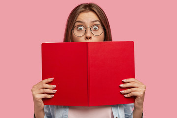 Photo of intelligent female student covers face with red book, stares at camera with bugged eyes,...