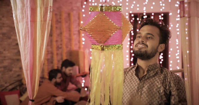 A smiling handsome man hanging a Diwali traditional lantern during the festival. A young male wearing kurta hanging colorful Diwali Kandil while his colleagues sitting in the background