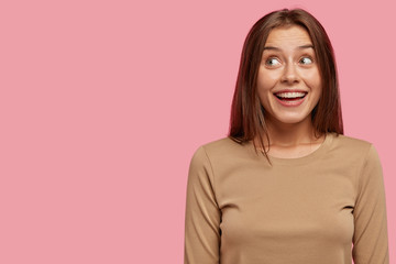 Fototapeta premium Studio shot of glad European young female has broad smile, looks positively and with dreamy expression aside, dressed in casual sweater, isolated over pink background with copy space on left