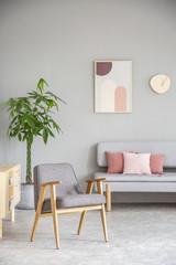 Wooden armchair in pink and grey living room interior with poster above sofa with pillows. Real photo