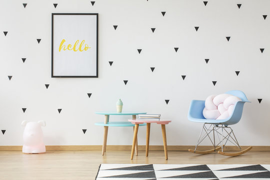 Fun wooden tables, light pink pillow in a baby blue rocking chair and a toy lamp in a cute nursery room interior with triangle stickers on a white wall