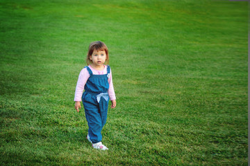 Fototapeta na wymiar Little Caucasian girl with an amazed face stands on a green meadow in a blue overall, copyspase