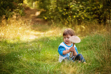A smiling girl of two years is sitting on the grass and holding an edible mushroom