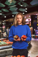 Fototapeta premium Young man in dark blue sweater thoughtfully looking on mangoes in hand in supermarket