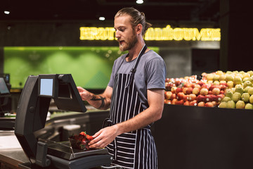 Young smiling seller in apron weighing strawberries on digital scales in supermarket