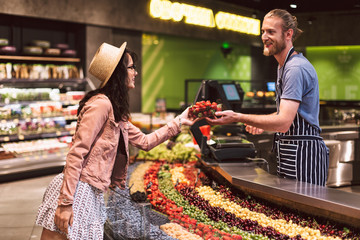 Obraz premium Young smiling seller in apron behind counter happily giving strawberries to customer in supermarket
