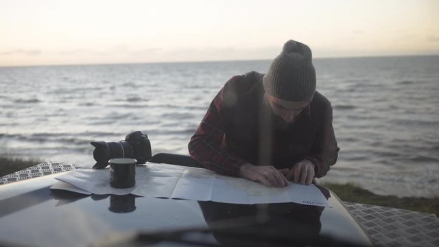 Outdoorsy man with a beard studying map on a bonnet of a vintage 4x4 by the sea, handheld