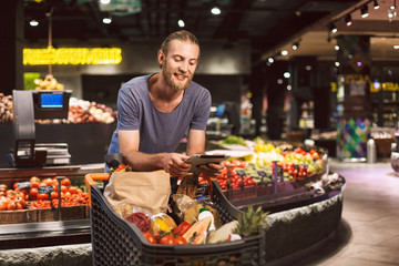 Young smiling guy leaning on trolley full of products while happily using the tablet in modern supermarket
