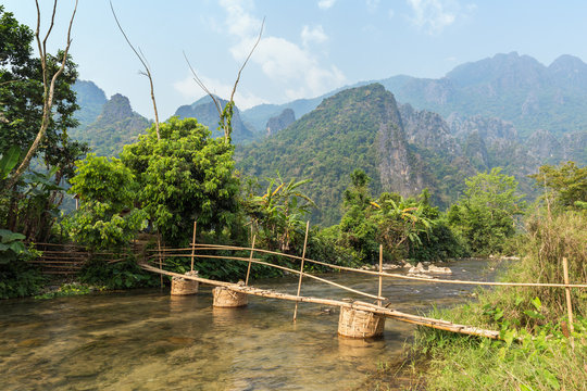 Beautiful view of a tiny bamboo bridge over a shallow river and striking karst limestone mountains near Vang Vieng, Vientiane Province, Laos, on a sunny day.