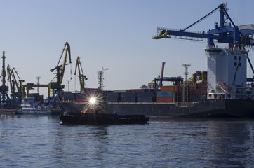 Fototapeta premium The tug floats and its spotlight emits beautiful rays in the background port cranes produce loading and unloading
