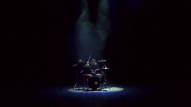 Drummer of a rock band on the stage plays drums during a musical performance, live show, indie rock, pop rock. Man playing drums on black background with smoke. Wide shot.