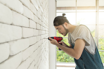 Young Asian man using electric drill on white brick wall in room, concept for home improvment, DIY...