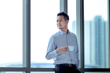 Fototapeta premium Young Asian businessman standing against window relaxed in his hands is cup of coffee look ahead.