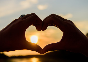 Fototapeta na wymiar Sunset sunlight romantic atmosphere. Male hands in heart shape gesture symbol of love and romance. Heart gesture in front of sunset above river water surface, defocused. Top places for romantic date
