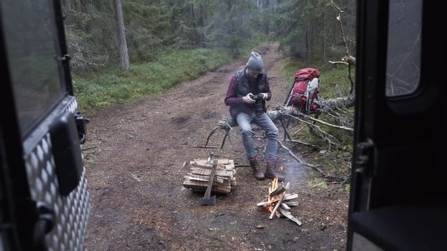 Outdoorsy photographer with a beard sitting by a camp-fire in forest and checking camera