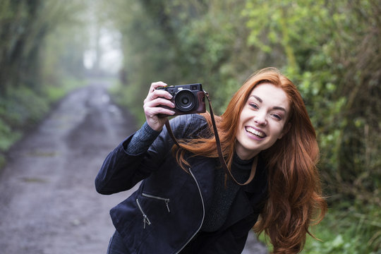 Smiling young woman with long red hair walking along forest path, taking pictures with vintage camera.