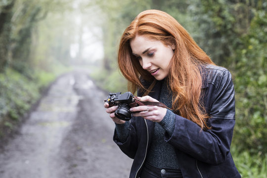 Smiling young woman with long red hair walking along forest path, taking pictures with vintage camera.