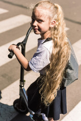 high angle view of beautiful little schoolgirl with long curly hair riding scooter and looking at camera