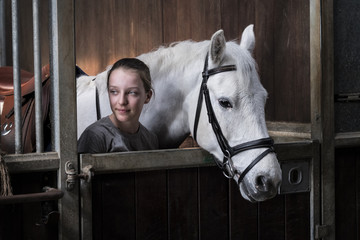 Teenage girl horserider with a grey horse outside astable, adjusting the girth and saddle.