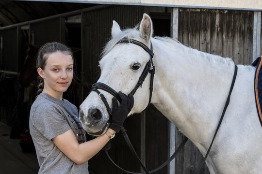 Teenage girl horserider with a grey horse outside astable, adjusting the girth and saddle.