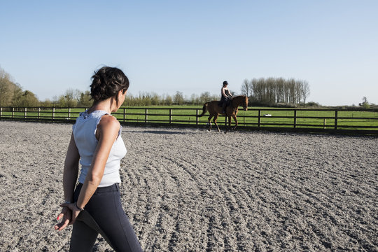 Woman trainer watching a teenage girl riding on a baybrown horse in paddock.