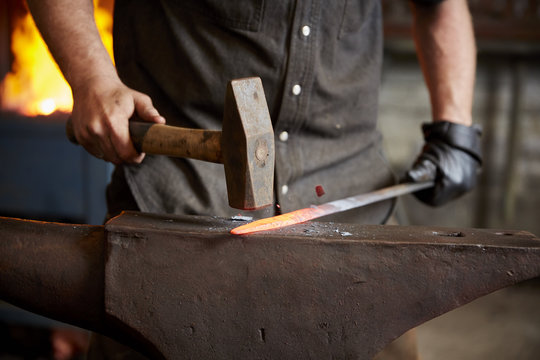 An artisan metal worker in ear protectors using a hammer to shape a red hot piece of metal on an anvil. 