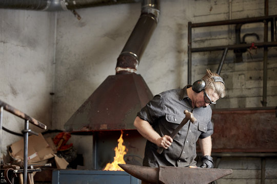 An artisan metal worker in ear protectors using a hammer to shape a red hot piece of metal on an anvil. 