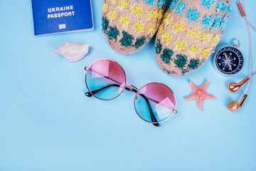 Travel flatlay with biometric passport, gradient round sunglasses, earphones, compass and espadrille sandals on the bright blue background. Decorated with sea shells and starfish.