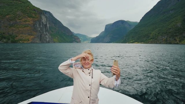 A happy woman takes pictures of herself on the nose of a cruise ship, sails through a picturesque fjord in Norway